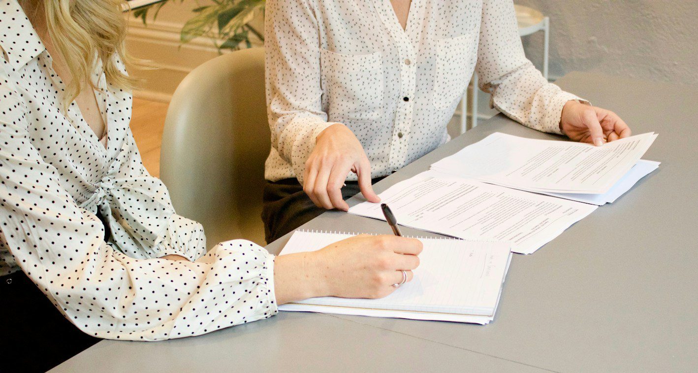 woman signing on white printer paper beside woman about to touch the documents